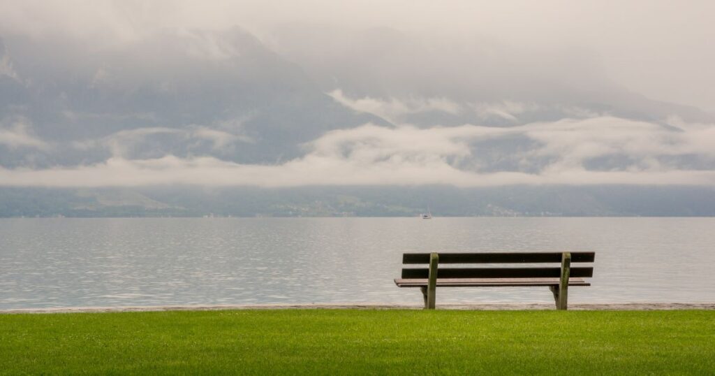 曇り空の湖畔に置かれた一人用ベンチ。転勤先での孤独や寂しさを表現した画像